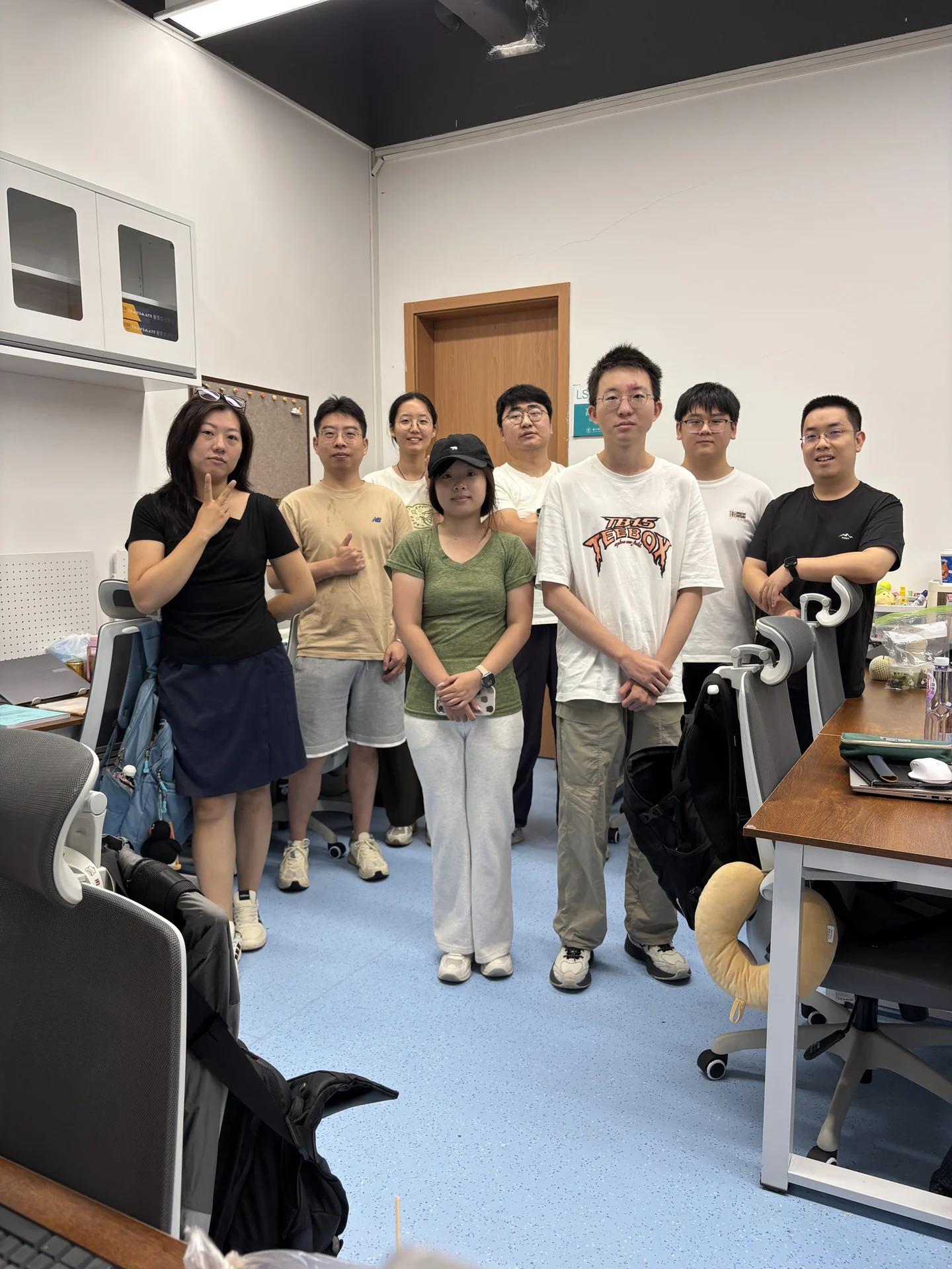 Eight group members standing together between desks in the lab office.